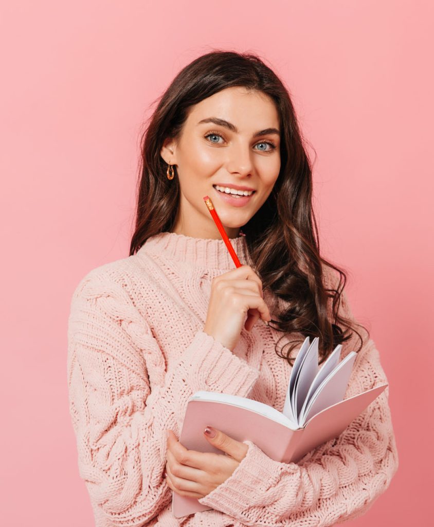 A young woman holding a notebook and pencil on a pink background.